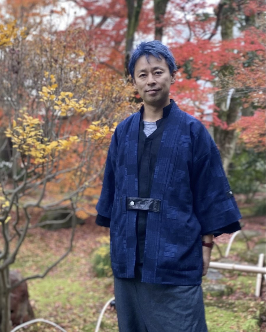 村松高志。紅葉の庭で、藍色の作務衣を着て立っている。